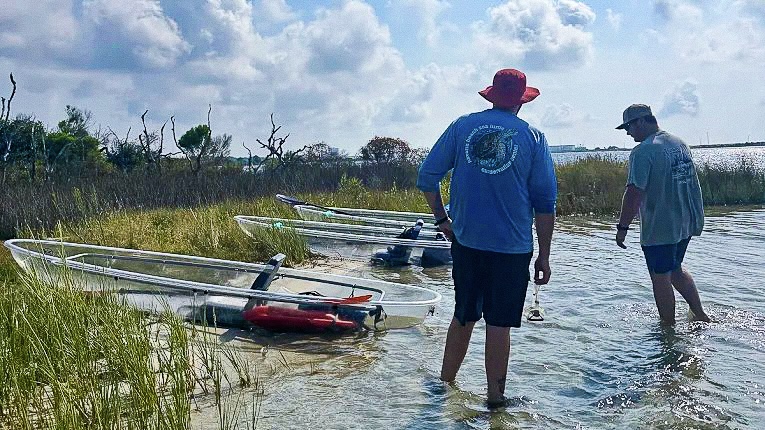 Two people in water near several beached kayaks, with grassy shoreline and cloudy sky in background.