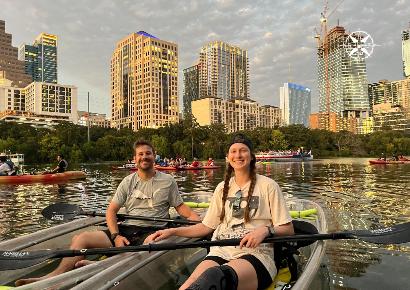 Two people kayaking on a river with city buildings in the background.