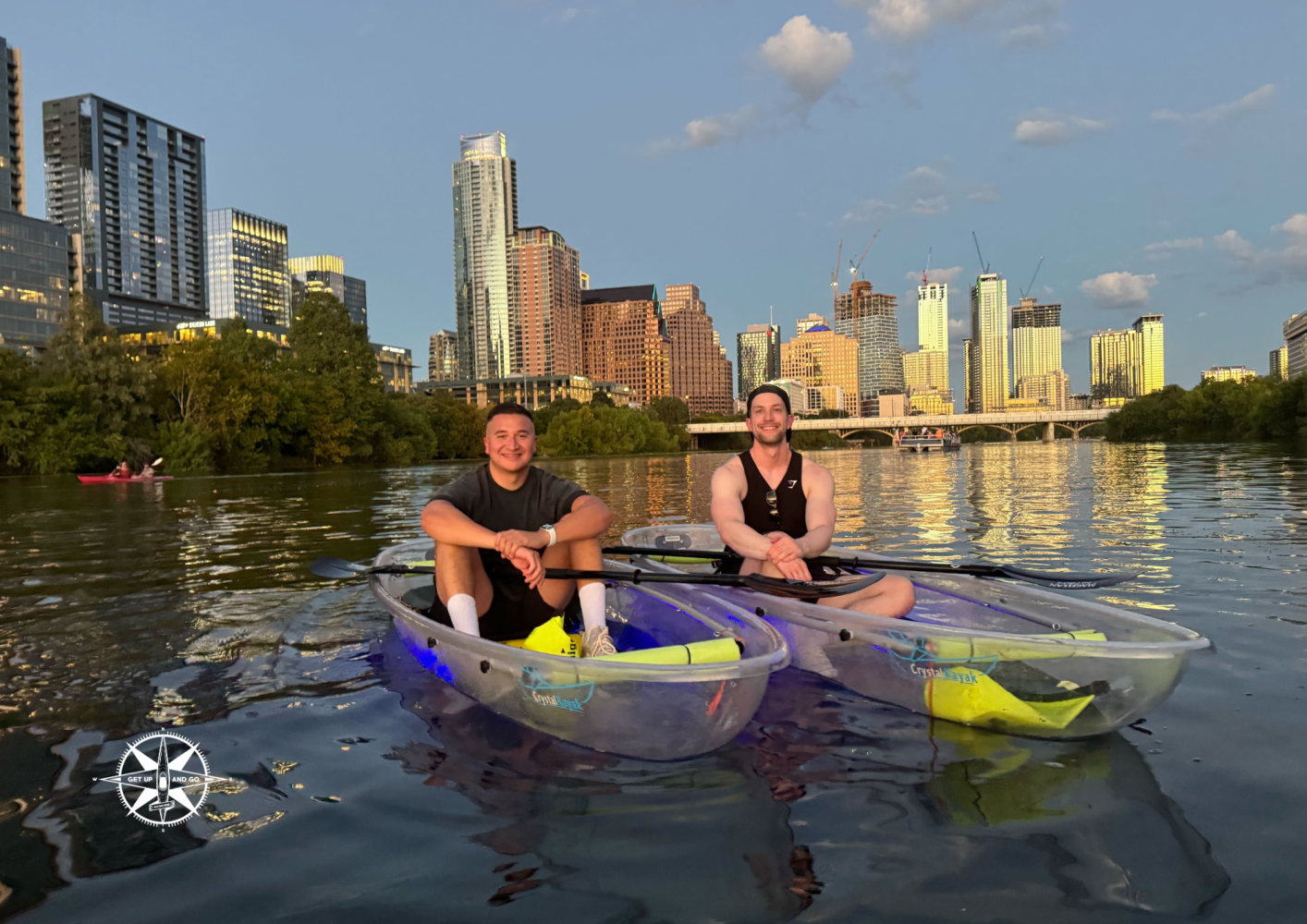Two people in clear kayaks on a lake with city skyline and trees in the background.