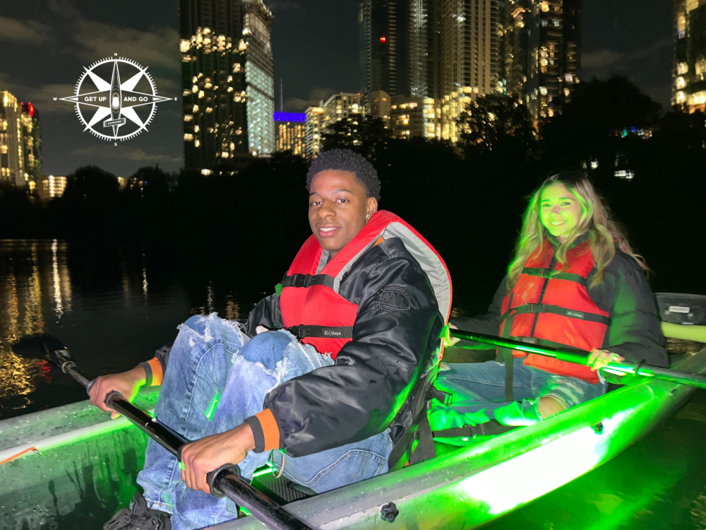 Two people kayaking at night with city lights in the background, wearing red life vests, green light illuminating kayak.