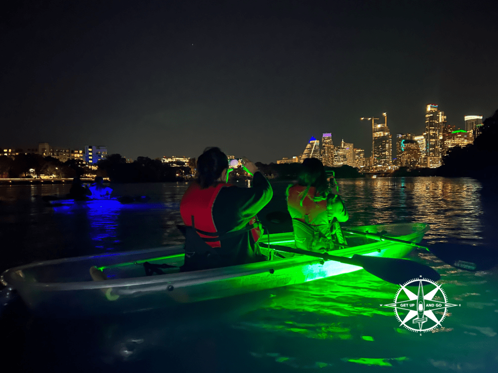 Kayakers with glowing boats on a river at night, city skyline in the background.