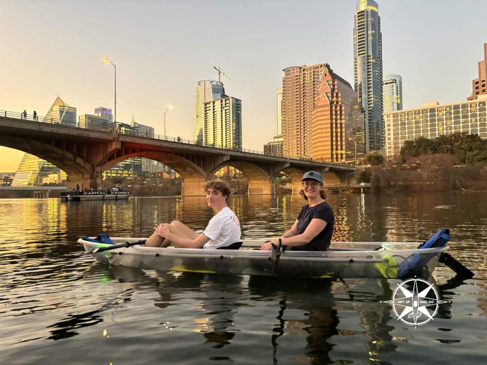 Two people kayaking on a river with a city skyline and bridge at sunset.