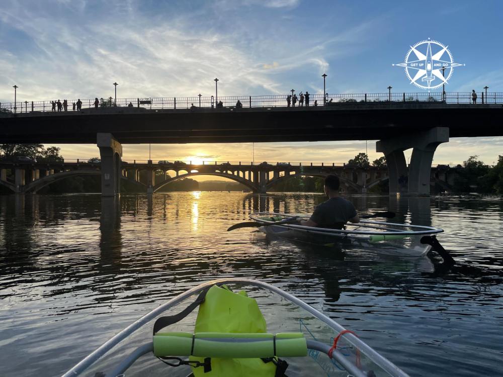 Two kayakers on a river at sunset under a bridge with silhouettes of people above.