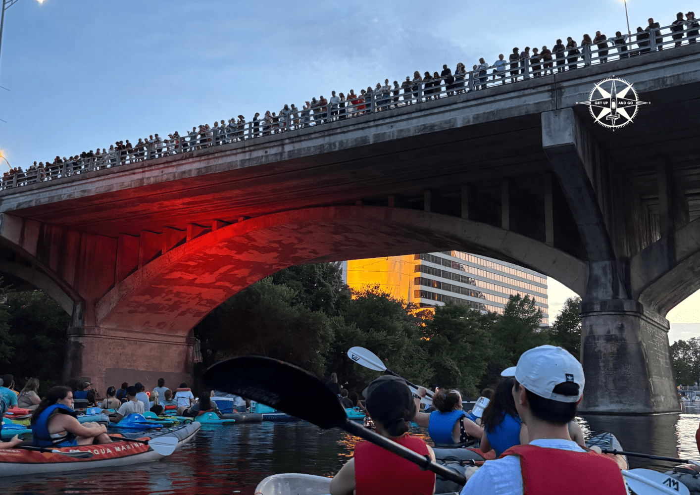 People kayaking under a bridge with spectators watching from above during sunset.