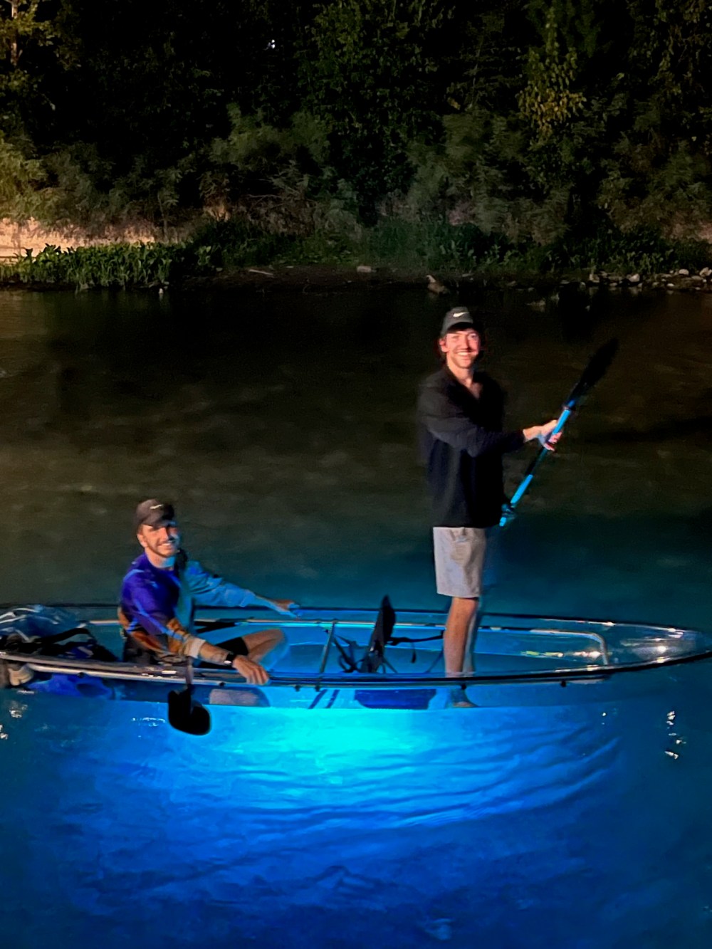 Two people on a transparent kayak with blue lights on a river at night.