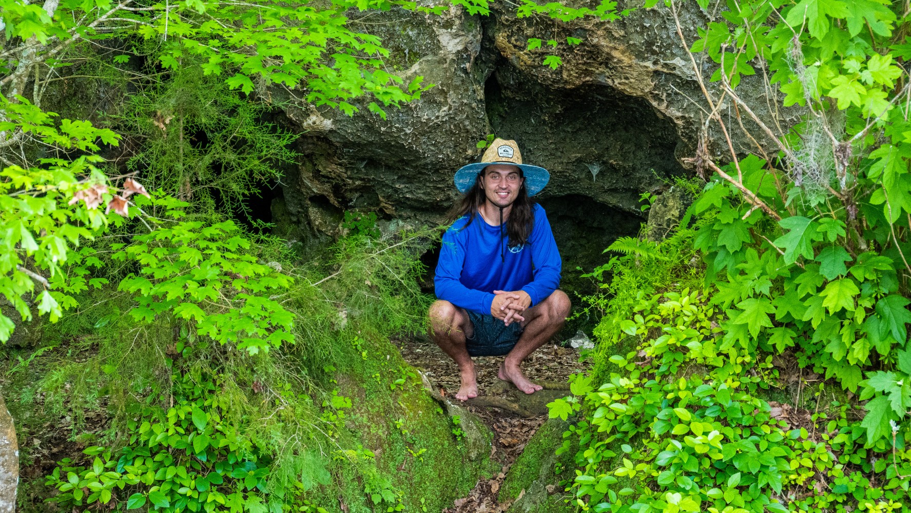 Person in blue shirt and straw hat crouching at a rock cave entrance surrounded by green foliage.