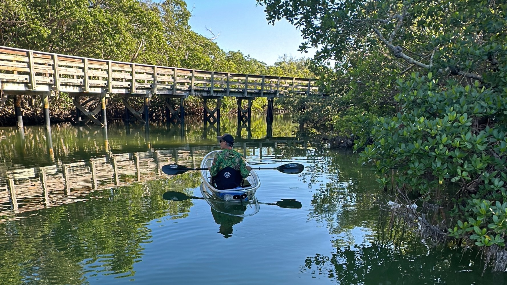 Person kayaking on a clear waterway beneath a wooden bridge, surrounded by lush greenery.