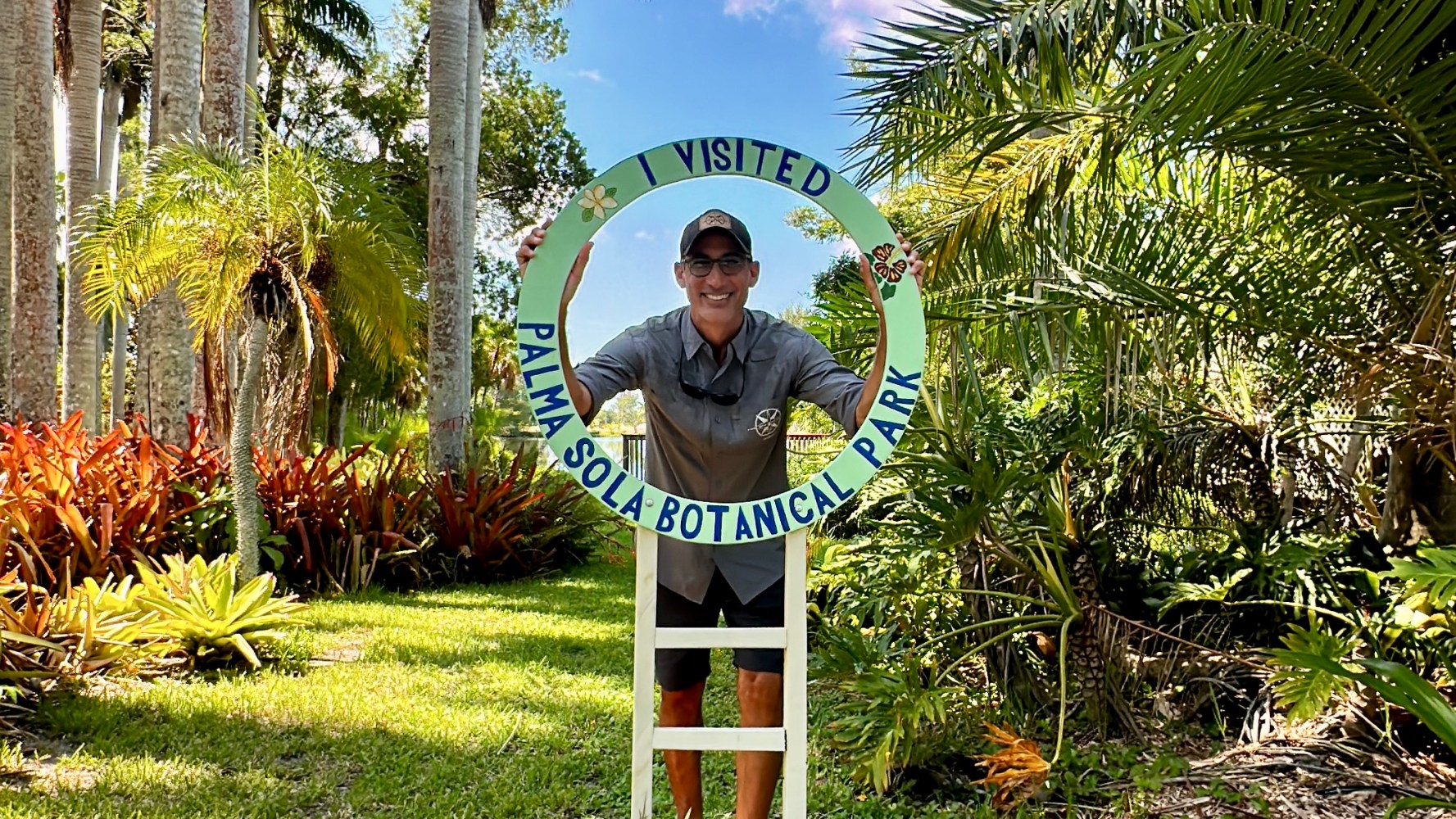 Person posing with a sign that says 'I Visited Palma Sola Botanical Park' amid lush green plants and trees.