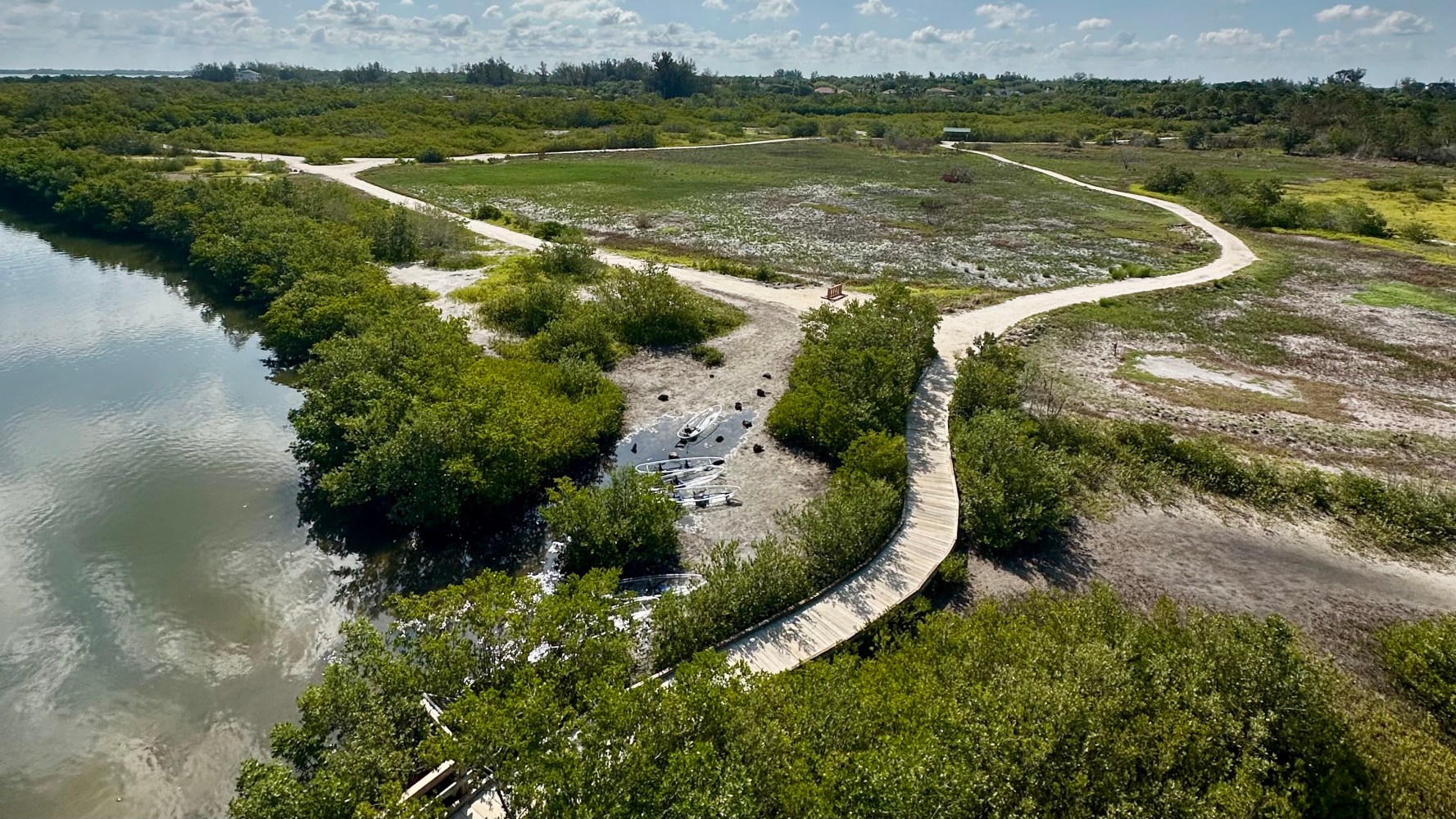 Aerial view of a winding boardwalk over green wetlands near a calm water body under a partly cloudy sky.
