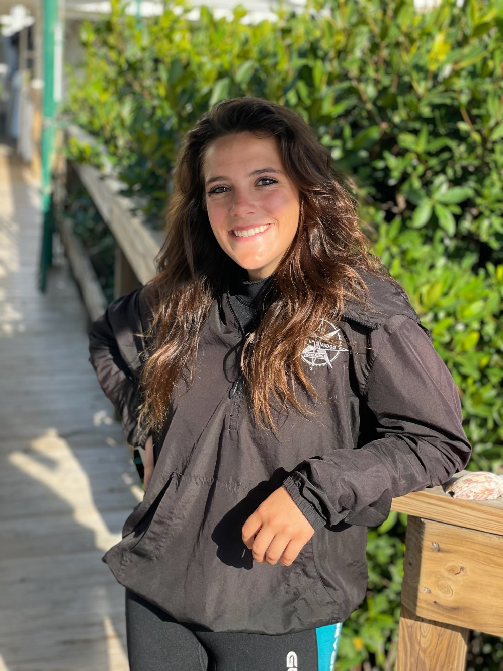 Smiling person in black jacket on a wooden path with green foliage.