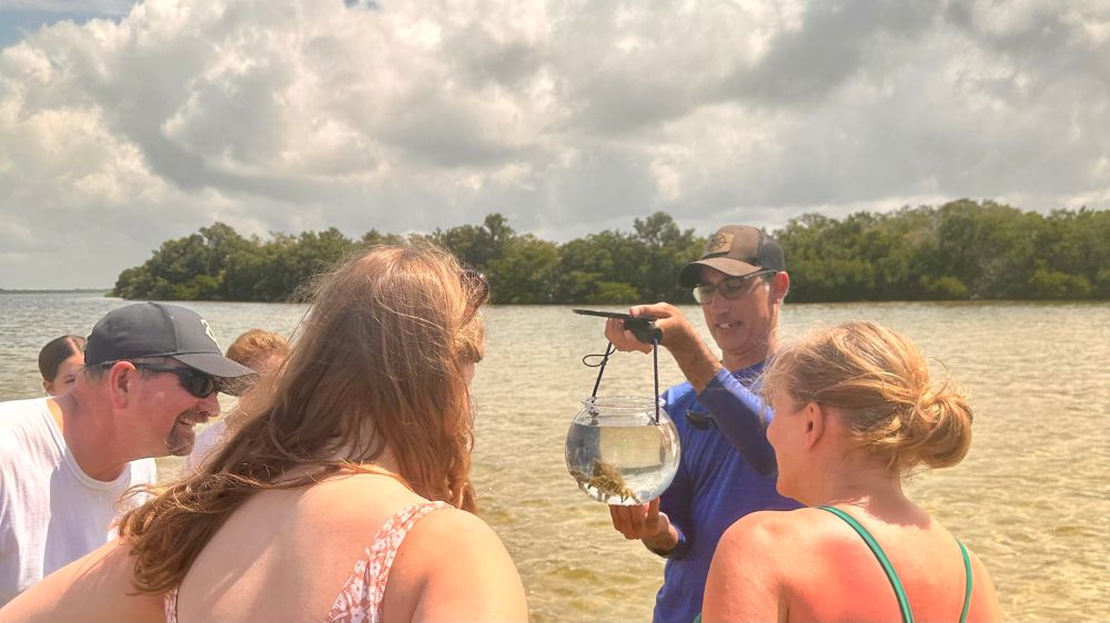 Group of people observing a man holding a small aquarium in a shallow water setting.