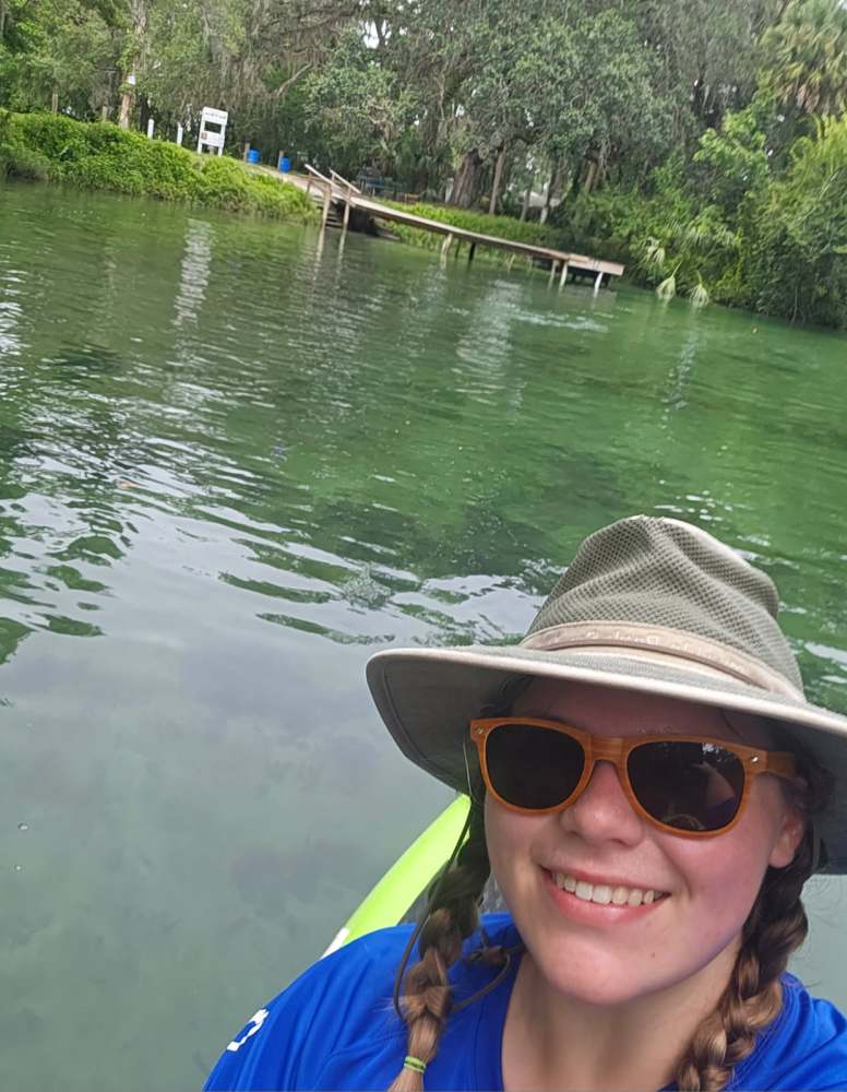 Person in hat and sunglasses kayaking on a river with green foliage and a small dock in the background.