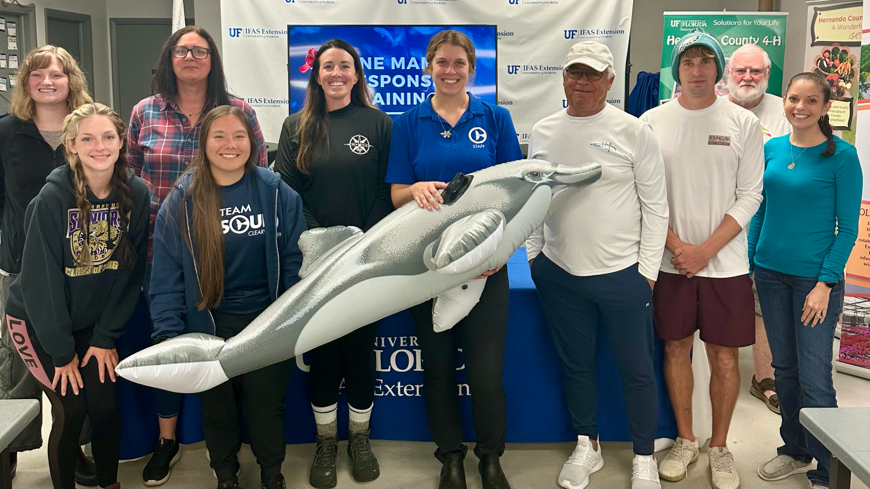 Group of people posing with a dolphin float at an event with banners in the background.