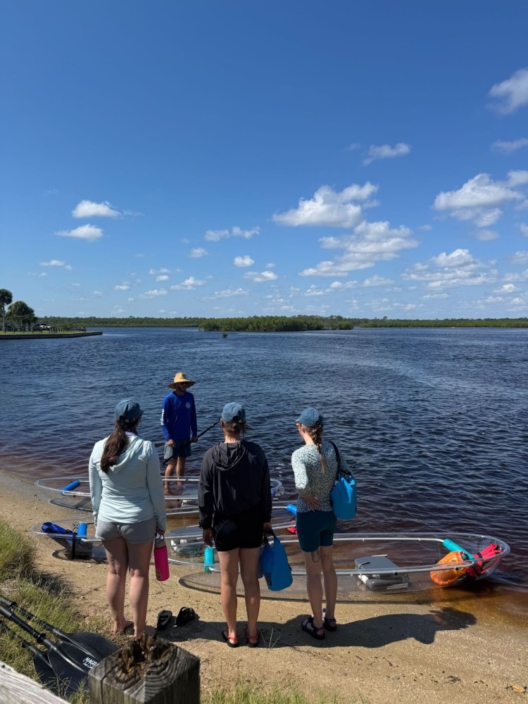 Four people with gear stand near clear kayaks on a riverbank under a blue sky.