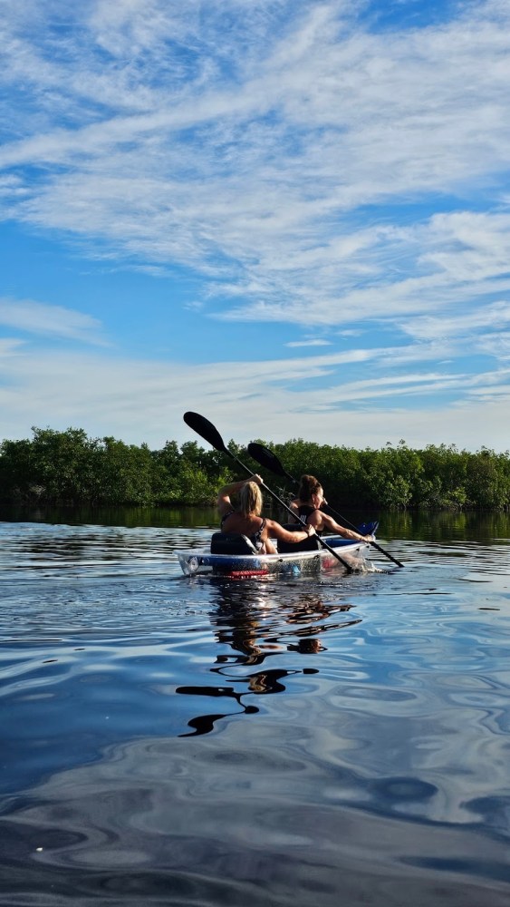 Two people kayaking on calm water under a blue sky with white clouds.