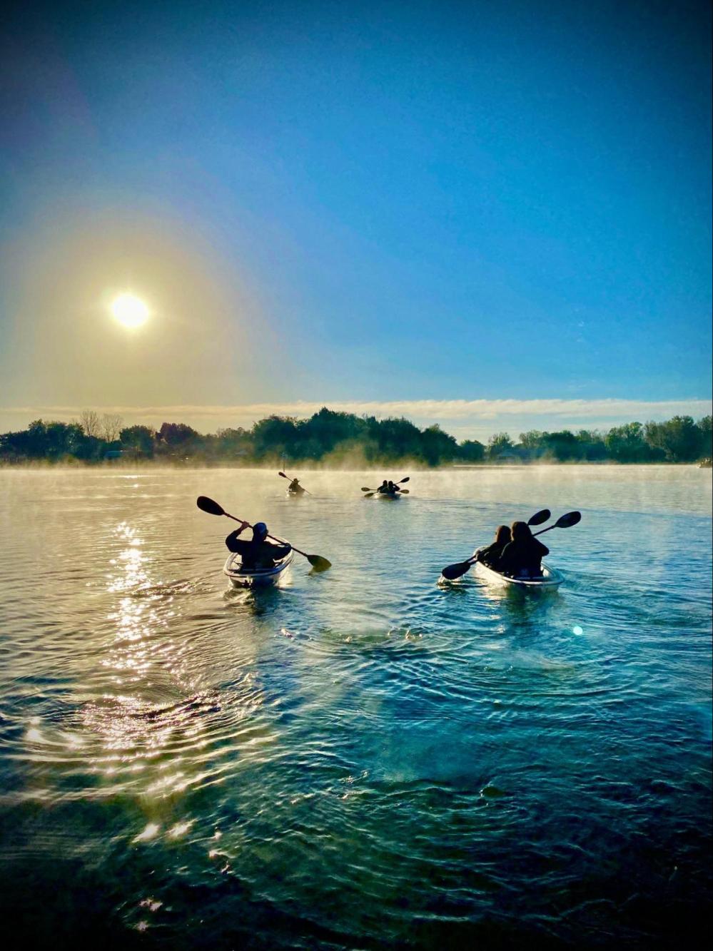 Kayakers paddle on a misty lake at sunrise, with trees in the background and a clear sky.
