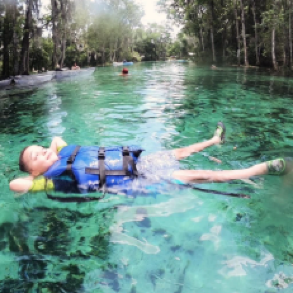Child in blue life jacket floats on clear water in a lush forest setting.