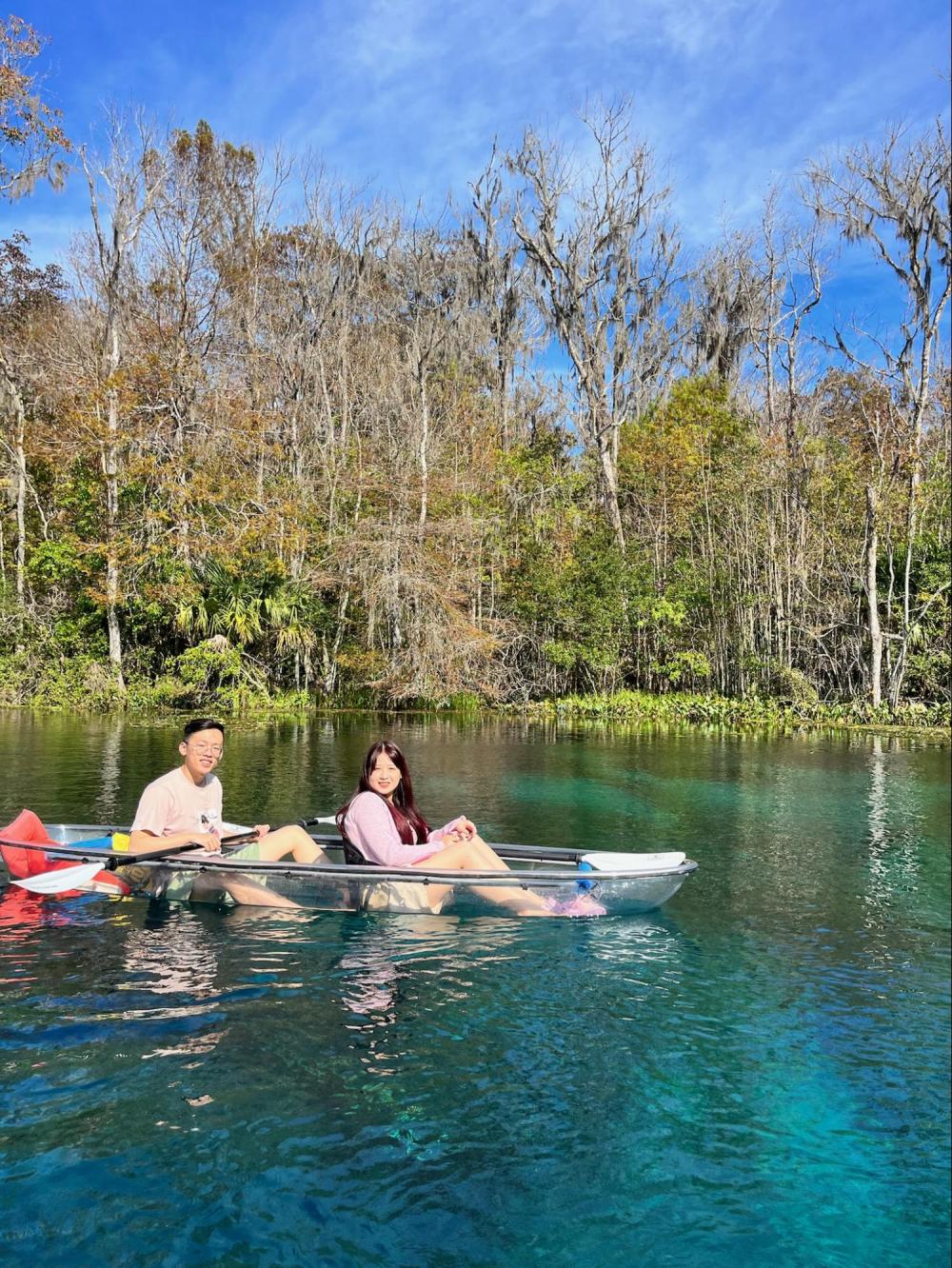 Two people kayaking on clear blue water near a forest.
