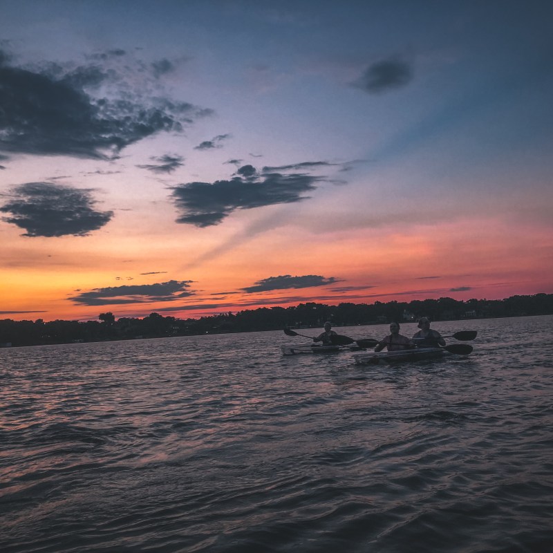 Three people kayaking on a lake at sunset with colorful sky.