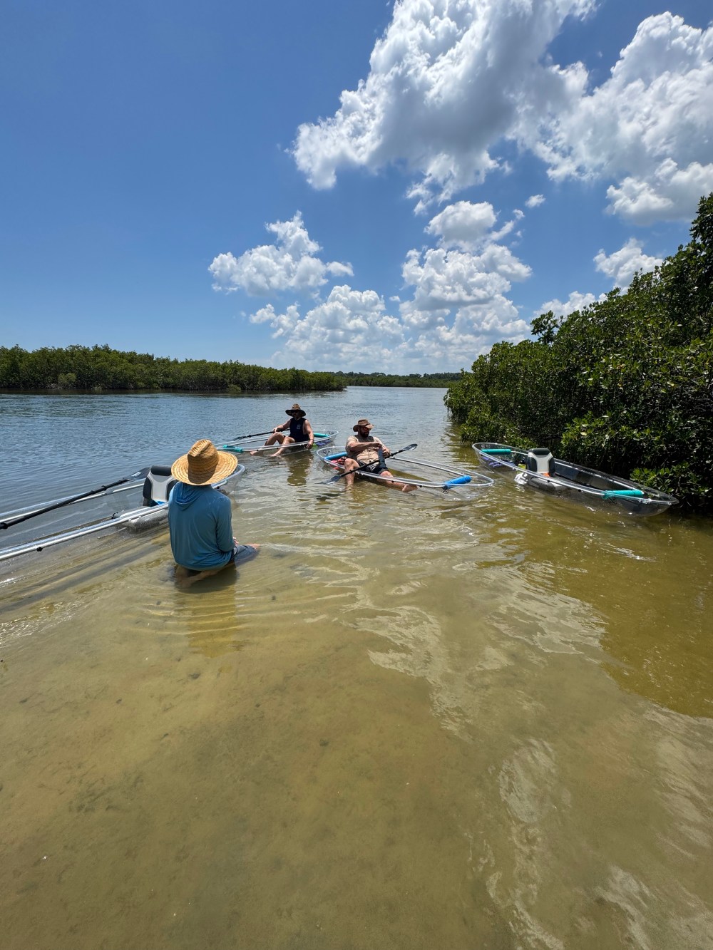 Three people on kayaks in a shallow river under a cloudy blue sky.