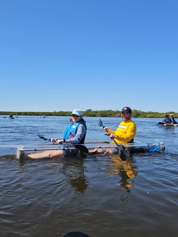 Two people kayaking in a see-through kayak on a sunny day with a clear blue sky.