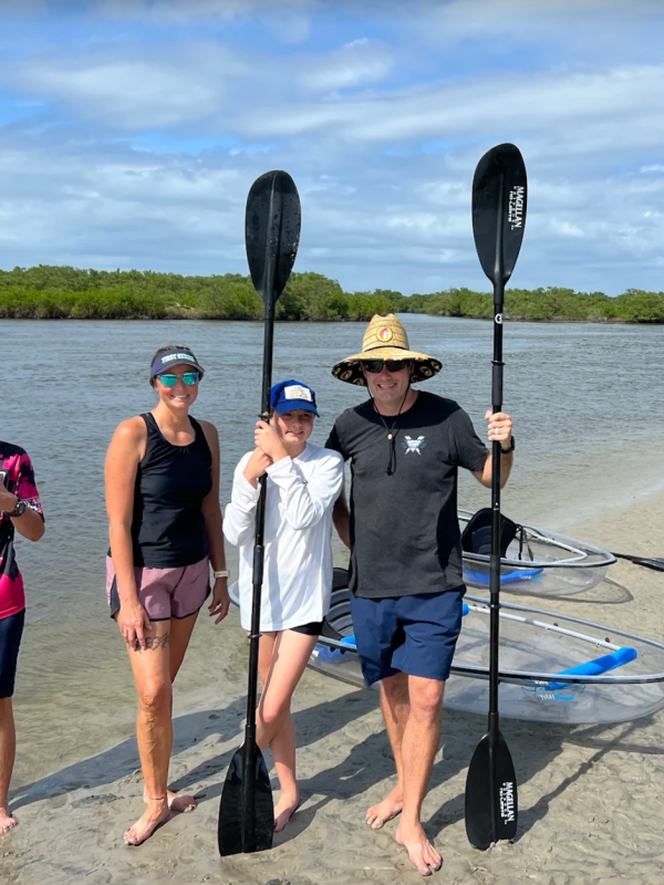 Four people with paddles standing on a sandy shore beside two clear kayaks, with water in the background.