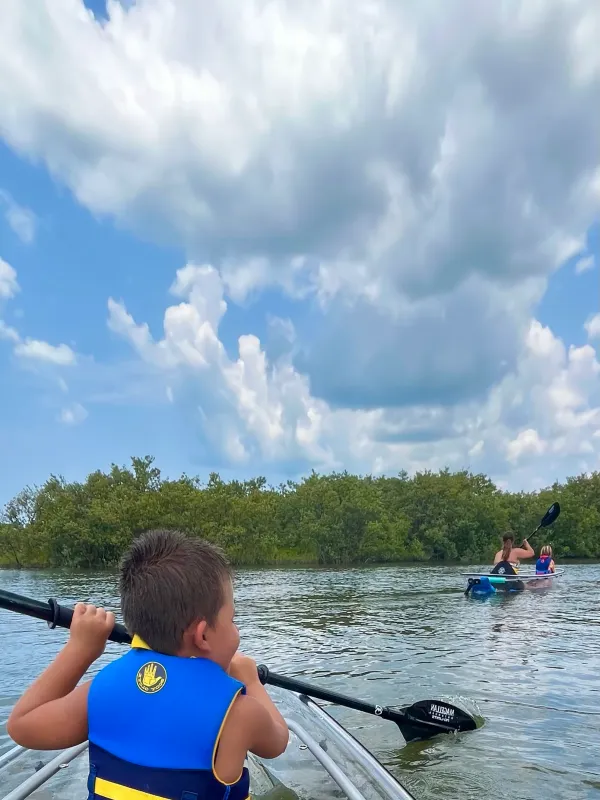 Child in a kayak with a paddle, wearing a life jacket under a cloudy sky.