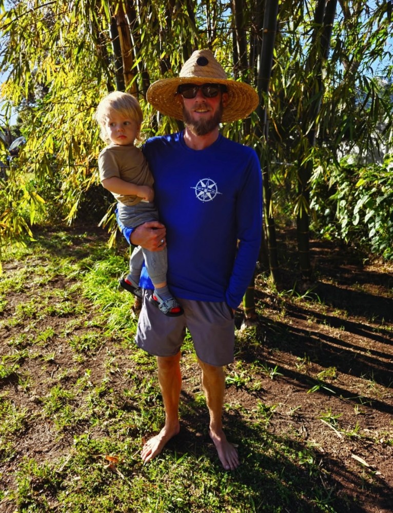 Man in hat and sunglasses holding child, standing barefoot on grass near bamboo.
