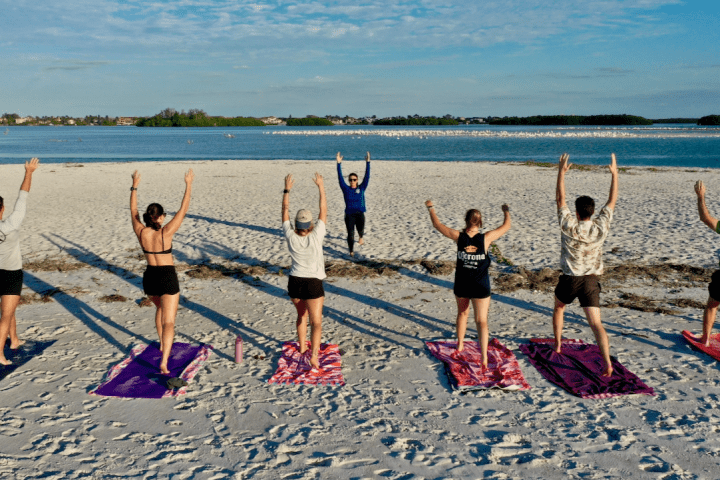 Seven people doing beach yoga, facing instructor and ocean, with towels on sand.