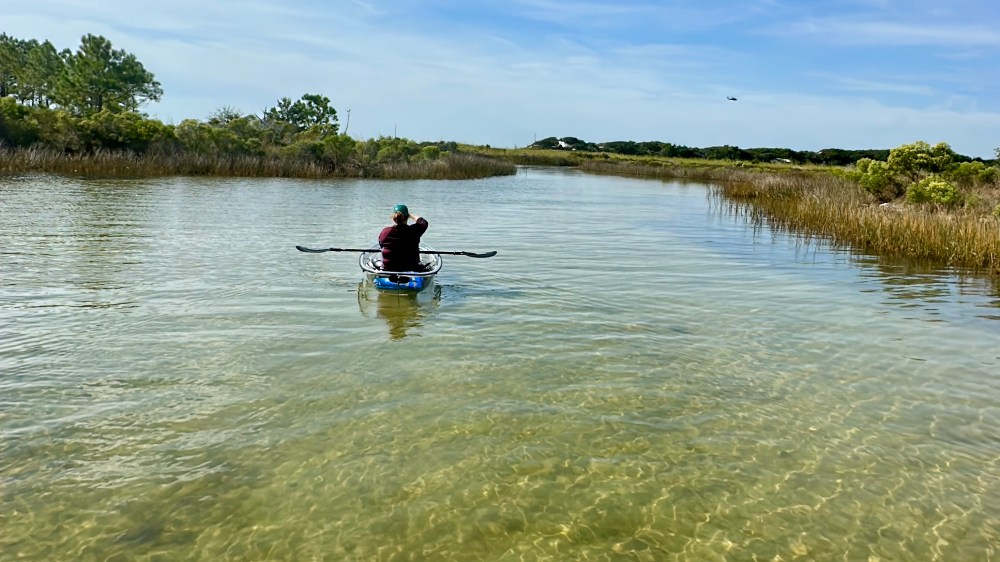 Person kayaking through a clear, shallow marshland under a blue sky.