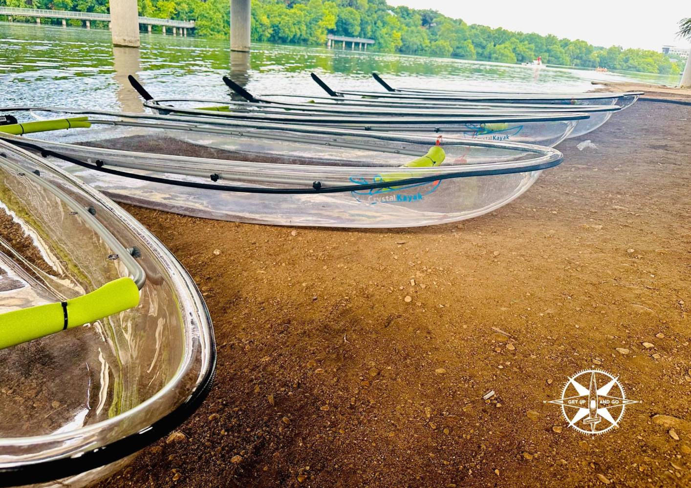 Clear kayaks with green paddles on a sandy riverbank, trees and water in the background.