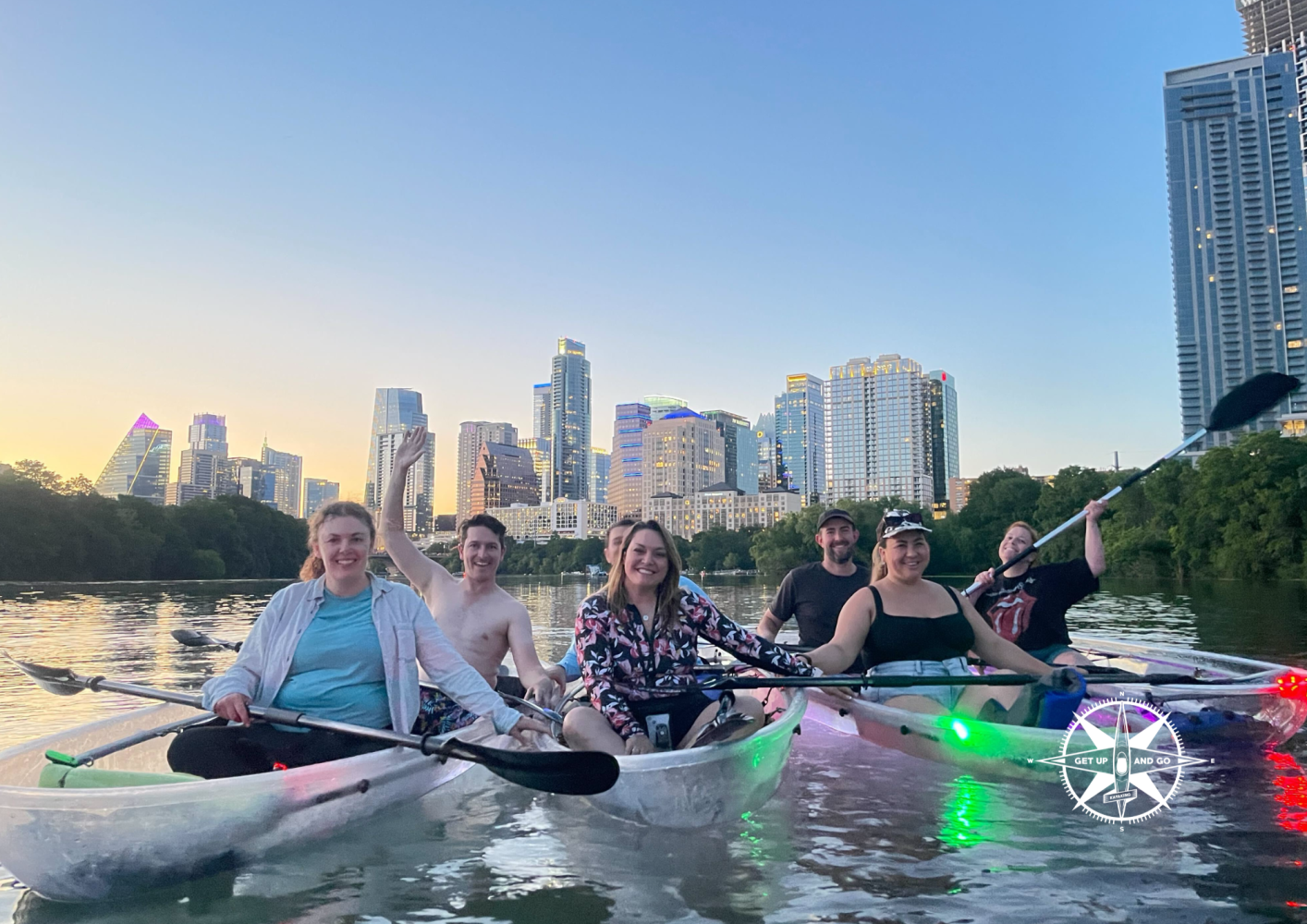Group of people in kayaks on a river with a city skyline at sunset.