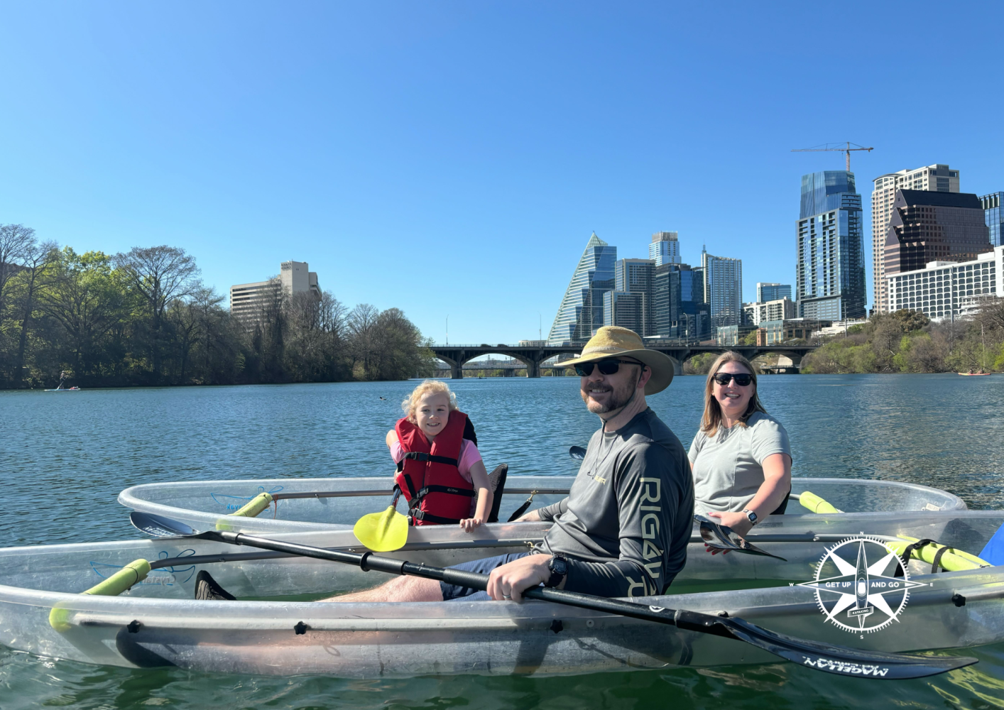 Three people kayaking on a river with a city skyline in the background.
