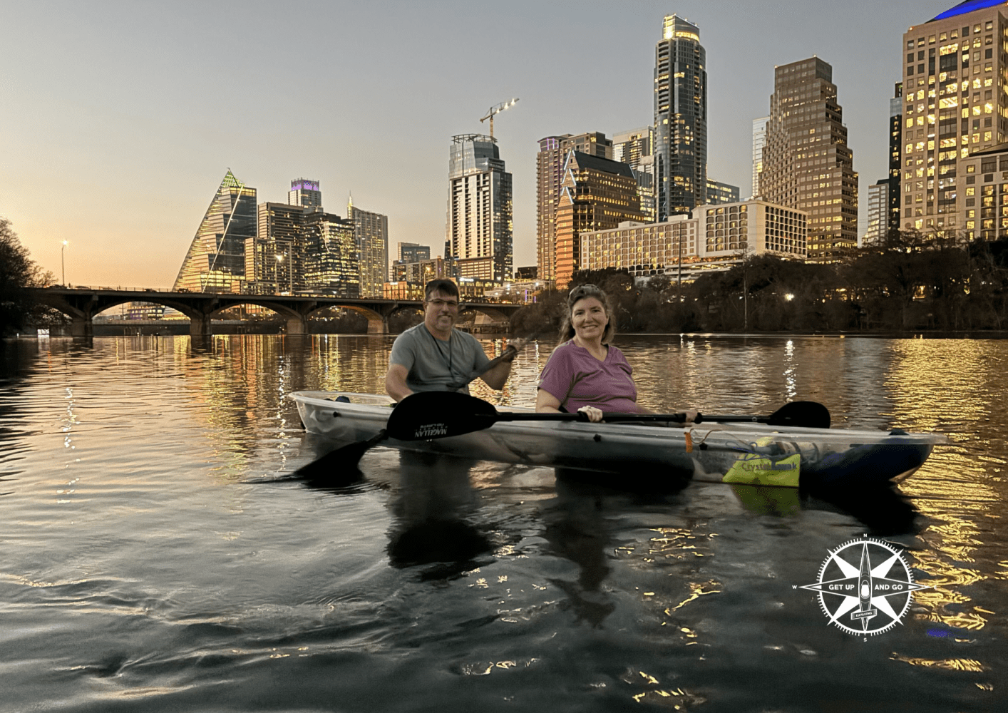 Two people kayaking on a river with city skyline at sunset.