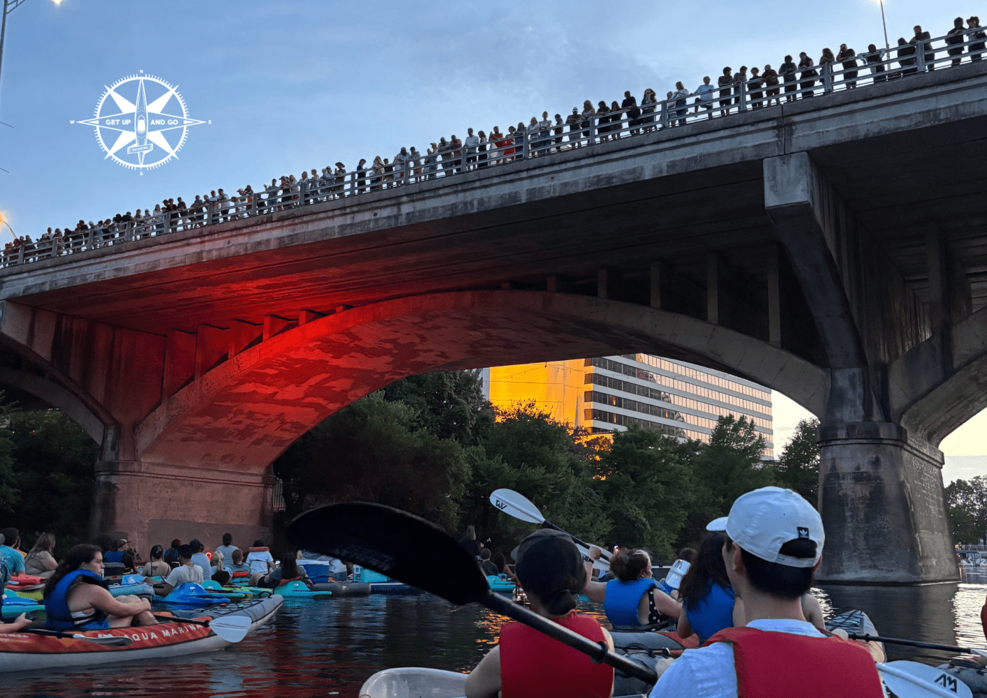 Kayakers on a river under a bridge at sunset, with people watching from above.