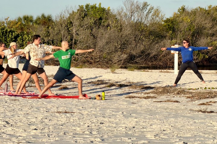 Group practicing yoga on a sandy beach led by an instructor, trees in the background.