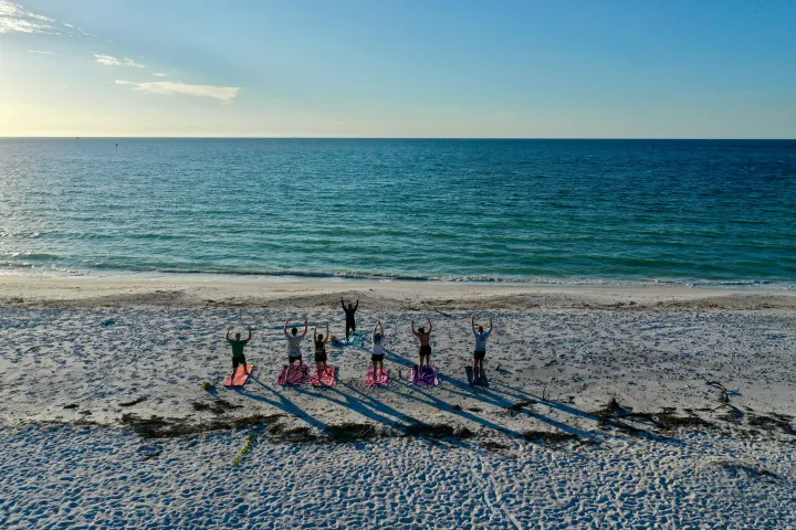 People doing yoga on a beach, facing the ocean, with long shadows on the sand.