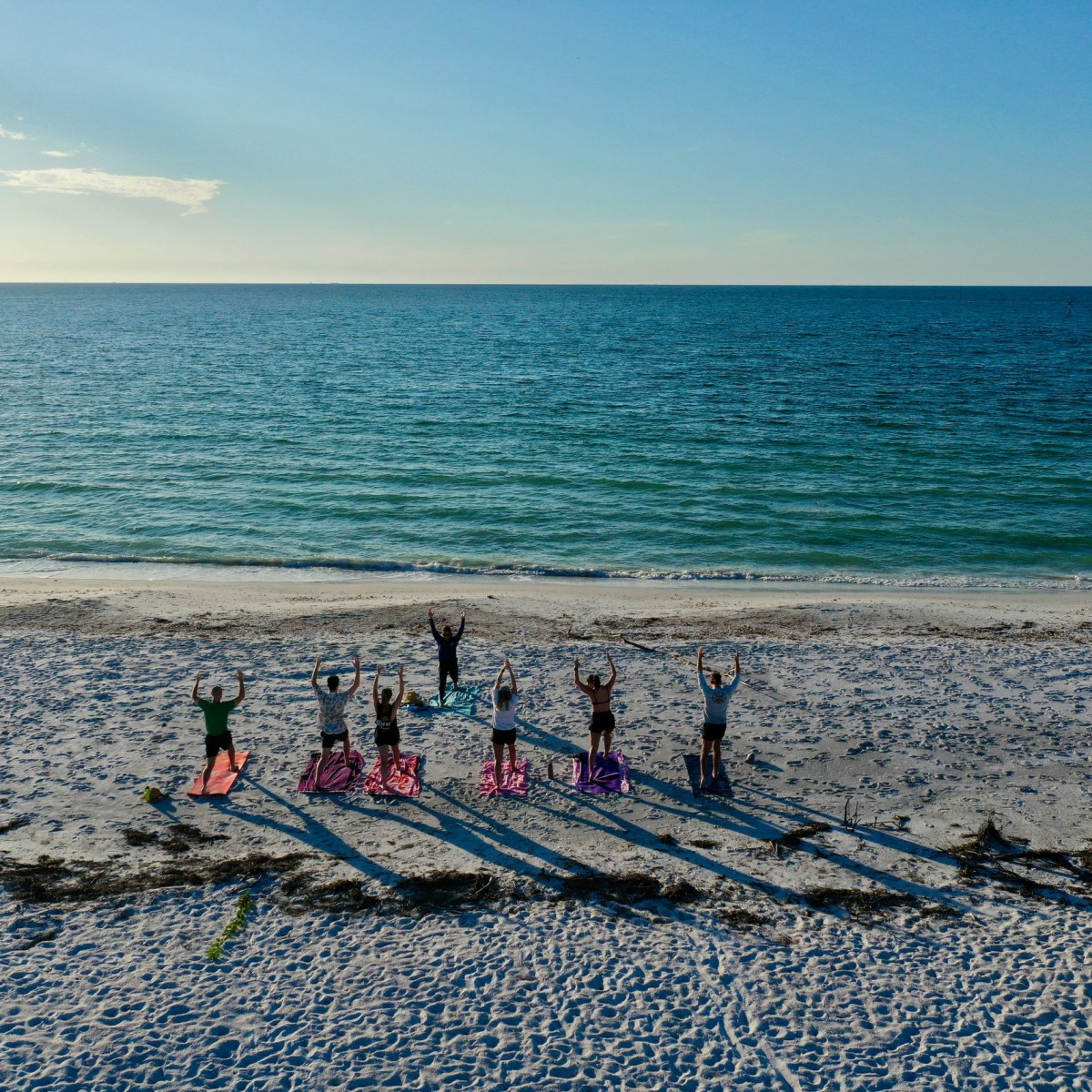 People doing yoga on a beach, facing the ocean, with long shadows on the sand.