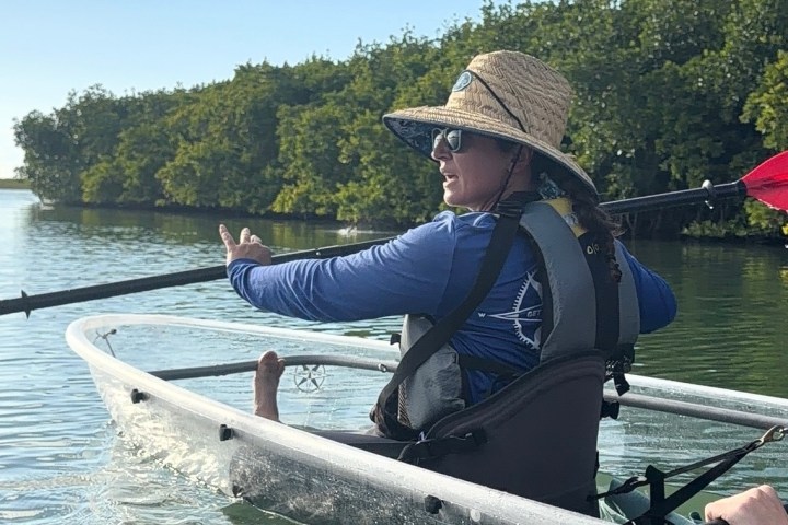 Person in a kayak with red paddles, wearing a sun hat and sunglasses, pointing towards trees.