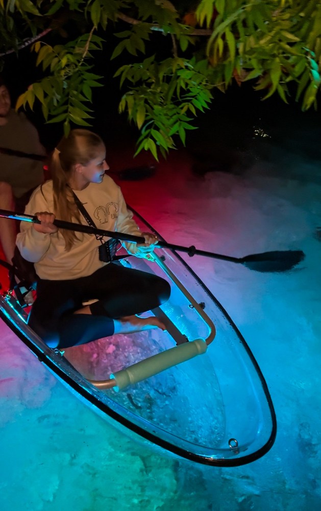 Woman kayaking at night in a transparent boat with colorful lights.
