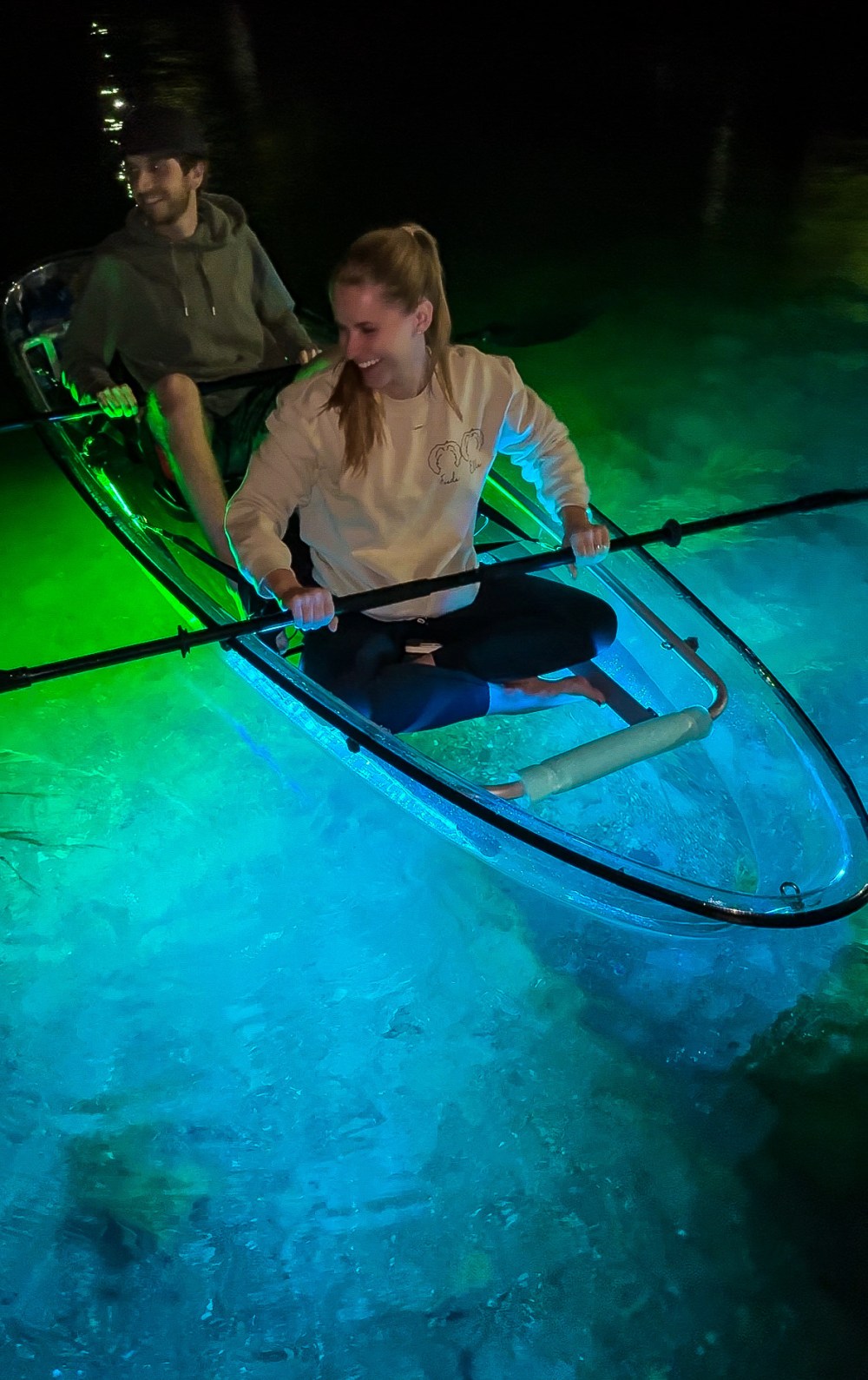 Two people in a transparent kayak glowing with green and blue lights at night.