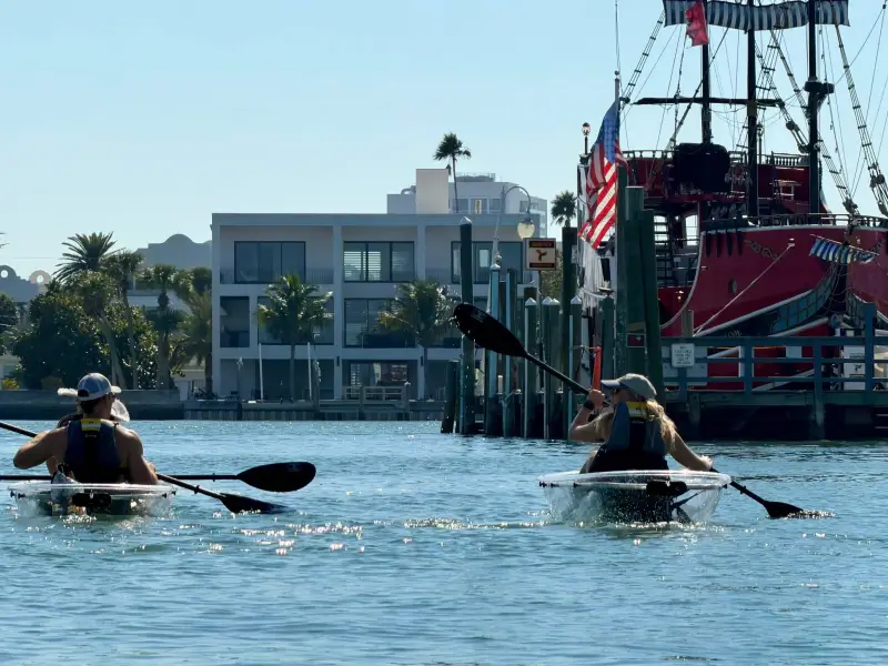 Two people kayaking near a docked pirate ship on a sunny day, with buildings and palm trees in the background.
