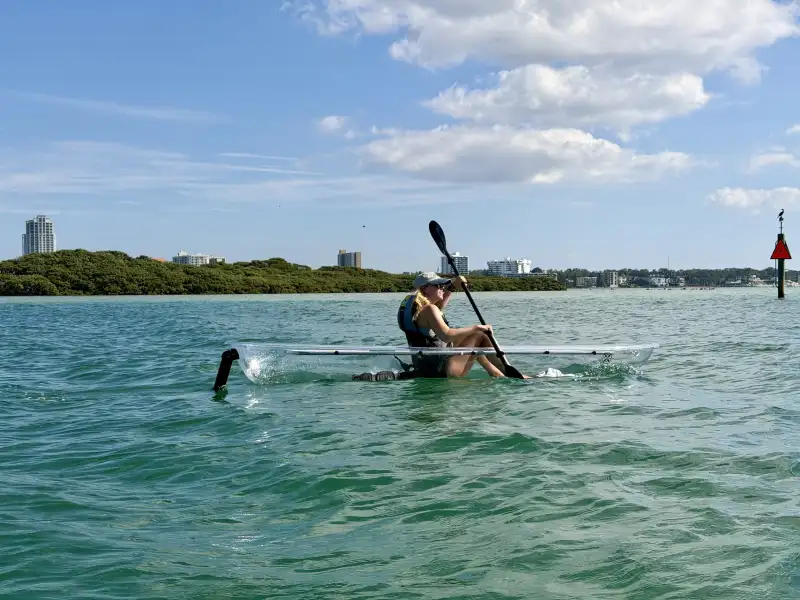 Person kayaking in a transparent kayak on blue water with city buildings in the background.