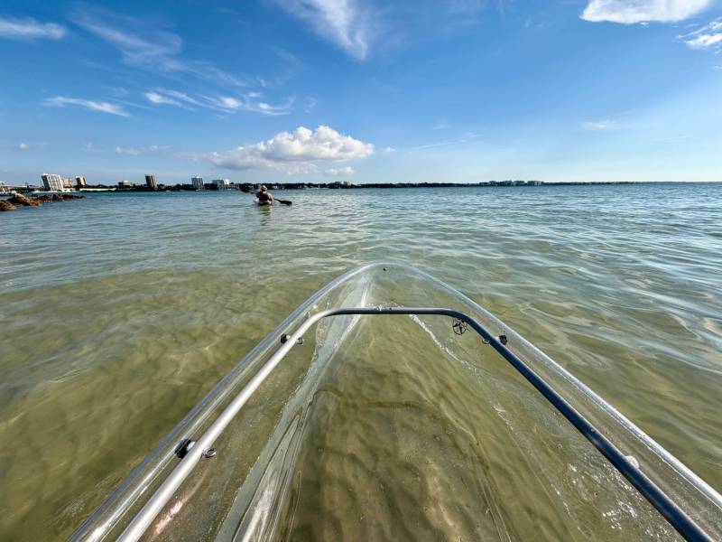 View from clear kayak on calm sea, with distant city skyline and blue sky.