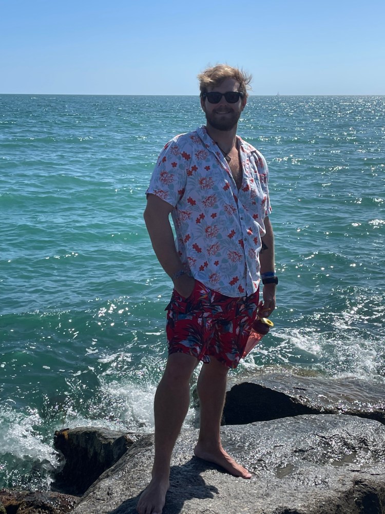 Man with floral shirt and shorts stands barefoot on rocks by the ocean, under a clear blue sky.