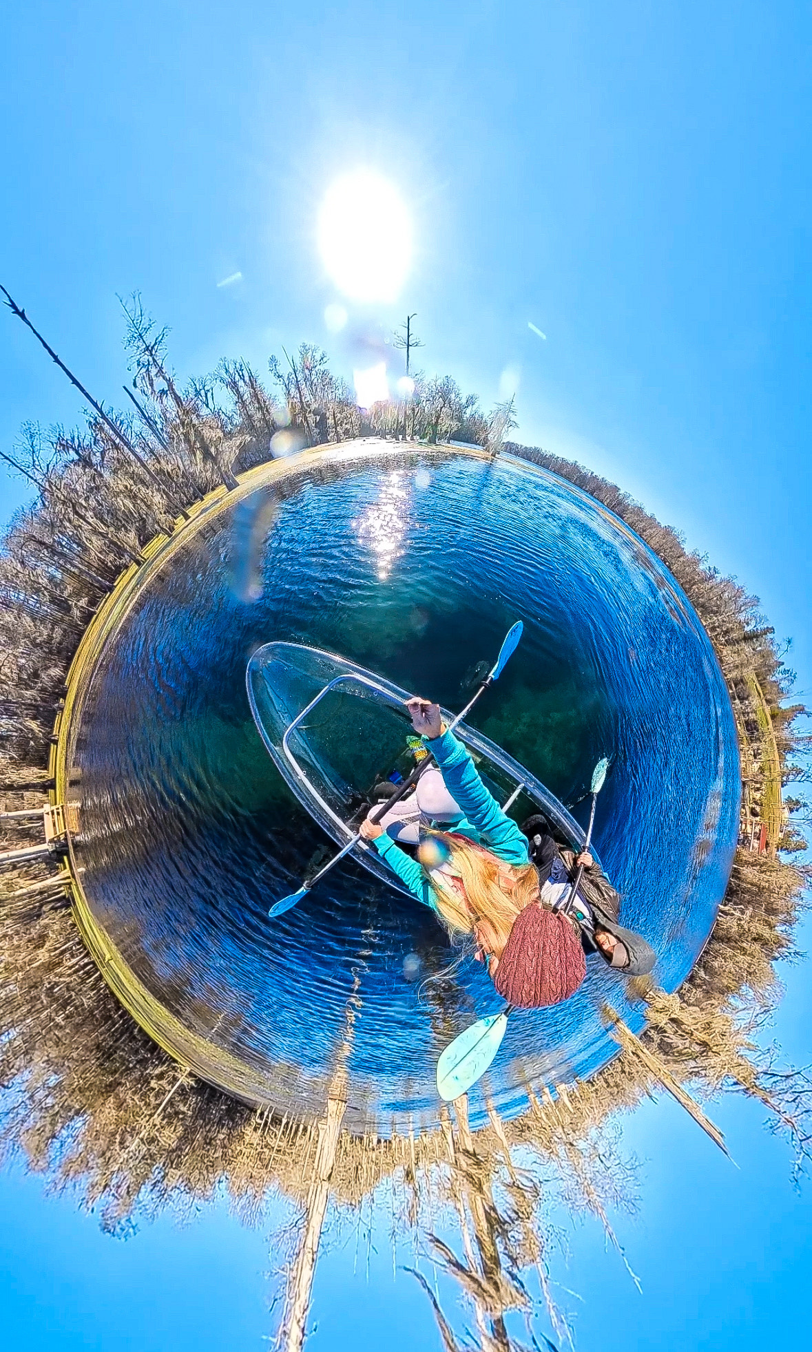 Two people kayak on clear water in a tiny planet view with sunny sky.