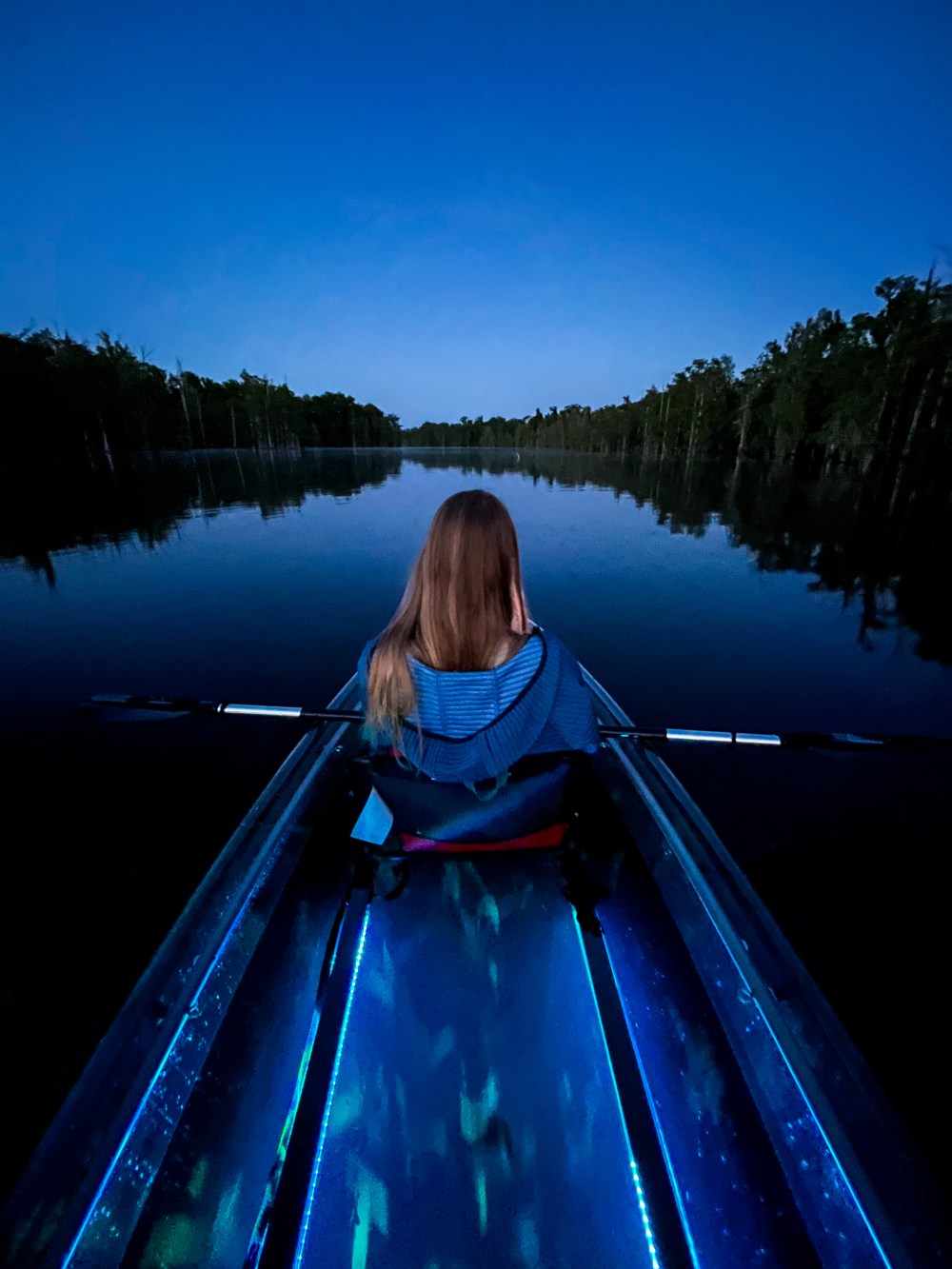 Person kayaking on a calm lake at dusk, with trees on the horizon.
