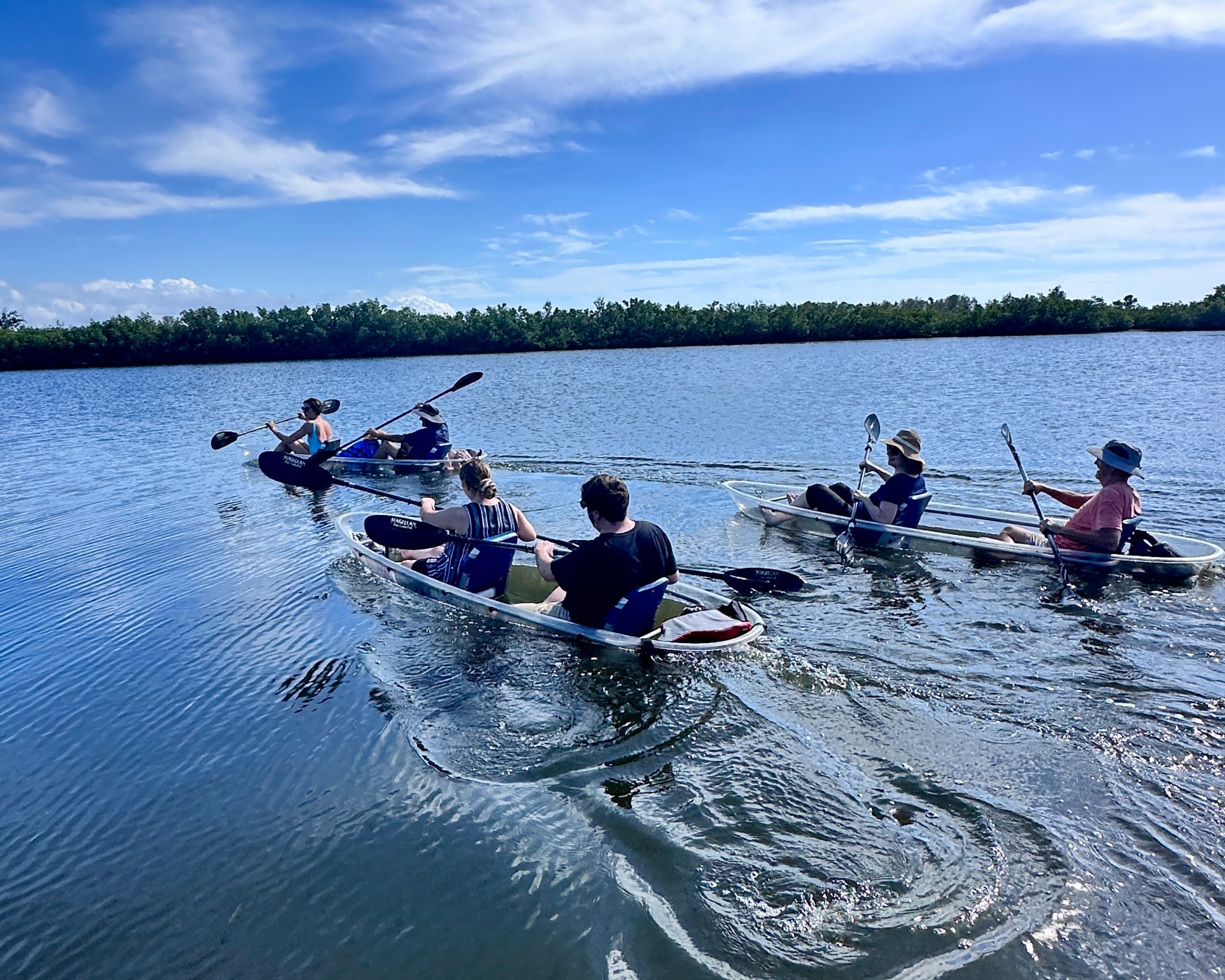 Five people kayaking on a calm lake under a blue sky with scattered clouds.