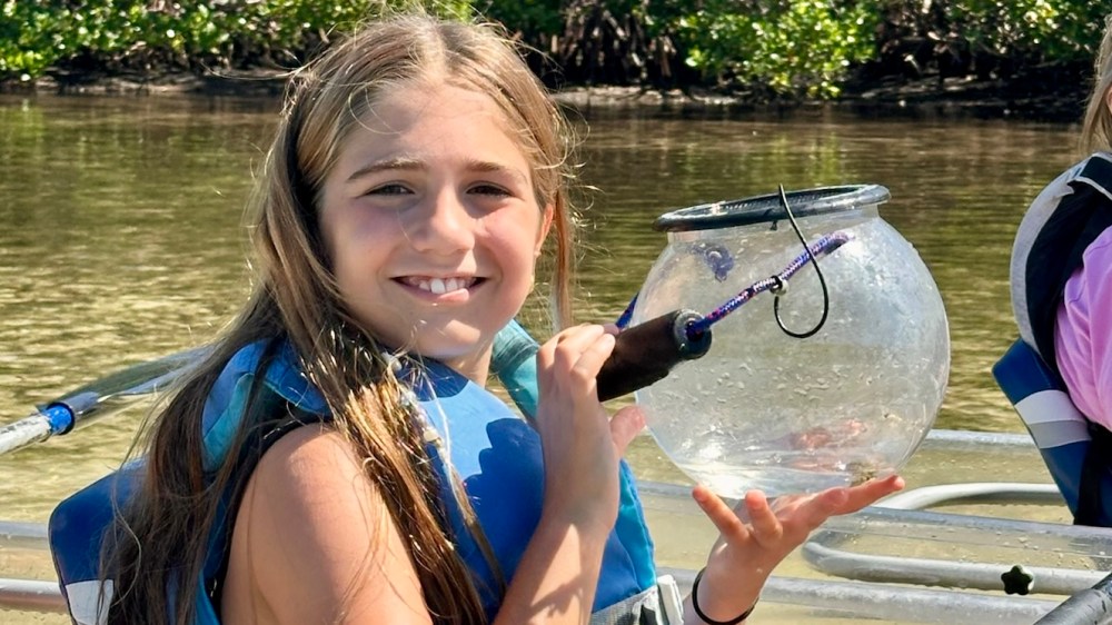 Girl smiling in a boat, holding a glass container with water, wearing a life jacket.