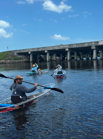 Three people kayaking under a bridge on a sunny day.